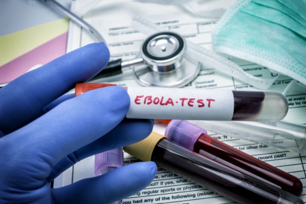A medically gloved hand holds a blood sample vial with the words "Ebola Test" written in red ink