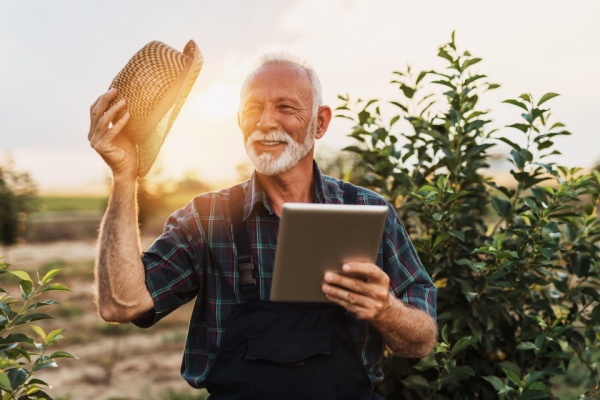 An elderly farmer stands in a field tipping his hat and holding a tablet