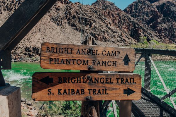 Carved wooden signs point to Bright Angel Camp, Phantom Ranch, and Bright Angel Trail in front of footbridge and Colorado River in Grand Canyon