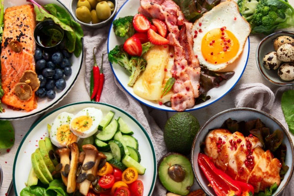 Plates filled with keto diet foods, including salmon, bacon, eggs, chicken, avocado and various fruits and vegetables, no grains