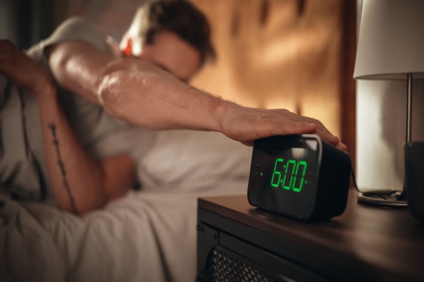 Man hitting snooze on alarm clock that reads 6:00AM