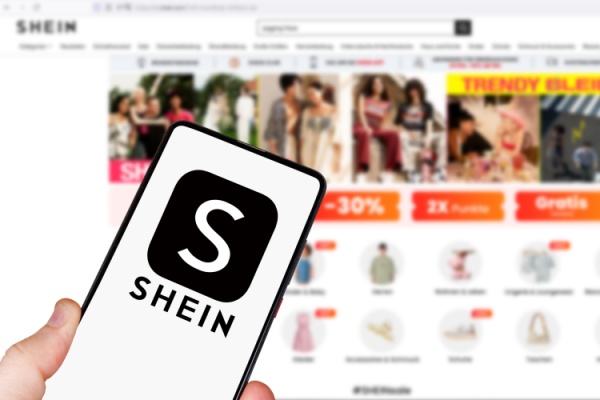Hand holds Shein logo and name on smartphone in front of computer display of website