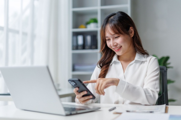 Young Asian woman working from home with open laptop and holding smartphone